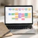 Laptop showing a colorful weekly calendar planner on screen with a notebook, pen and coffee on a white desk