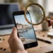 Hand holding a smartphone showing a photo, with a magnifying glass over the screen, laptop blurred in the background