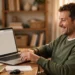 A smiling man at a tidy home desk closing old browser tabs on his laptop, feeling relieved after cleaning up his online accounts