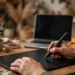 Hands drawing on a graphics tablet with a stylus at a warm, cozy home desk setup with a laptop and lamp